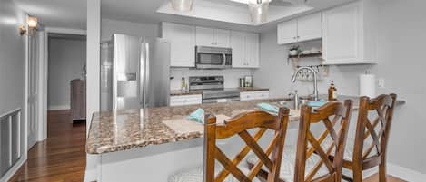 A kitchen with white cabinets, a granite countertop, a central island with chairs, stainless steel appliances, and pendant lighting.