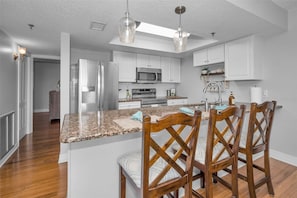 A kitchen with white cabinets, a granite countertop, a central island with chairs, stainless steel appliances, and pendant lighting.