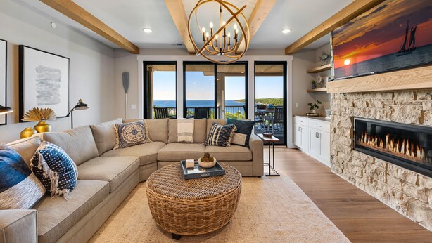 A modern living room with a sectional sofa, a round wicker ottoman, a fireplace, and a large window offering a view of the sea. - 302 with a View! Luxury 2 bed 2 bath condo with amazing views of Sister Bay (Sister Bay)
