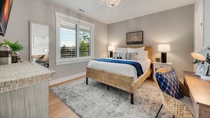 A bedroom with a bed, bedside tables, a chandelier, a window with blinds, and a view of greenery outside. - 302 with a View! Luxury 2 bed 2 bath condo with amazing views of Sister Bay (Sister Bay)