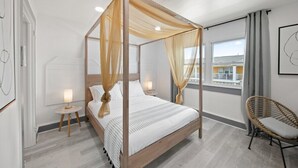 A four-poster bed with sheer curtains, a bedside table with a lamp, a wicker chair, and a view of buildings through a window.