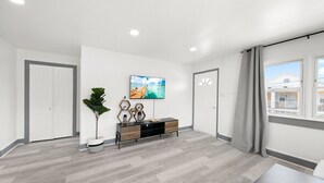 A spacious room with a grey wooden floor, a black and wood console table, a potted plant, a flat-screen TV mounted on the wall, and a window with a view of a building.