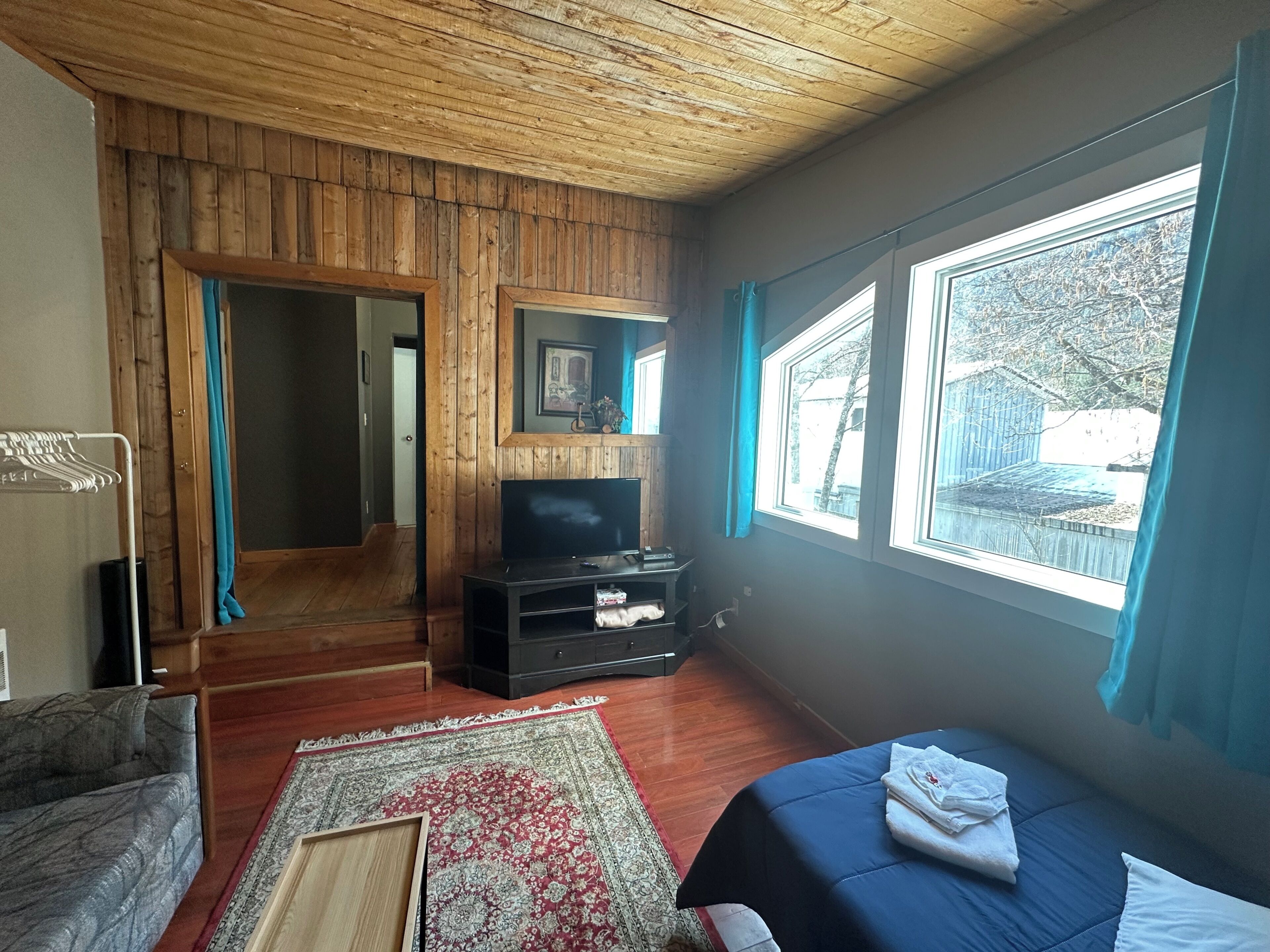 A room with a wooden ceiling, a television, a bed, a rug, and a window with curtains.