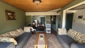 A living room with two grey sofas, a wooden coffee table, a bookshelf, and a kitchen in the background. - Historic Hotel Bayview (Stewart)