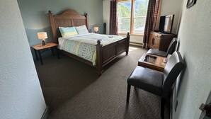 A bedroom with a wooden bed, a desk, and a chair. There is a window with curtains, a lamp on the nightstand, and a television on a cabinet. - Historic Hotel Bayview (Stewart)