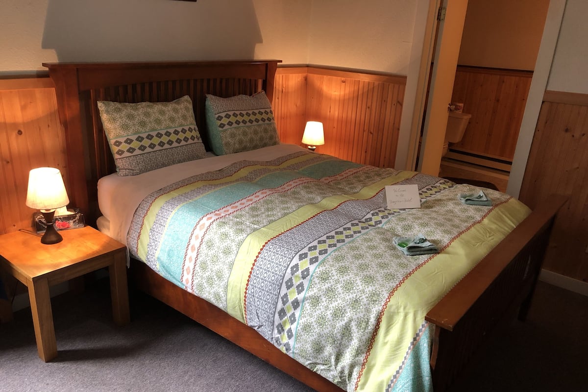 Una cama bien hecha con una colcha estampada, dos almohadas, una lámpara de noche y un cartel de bienvenida.