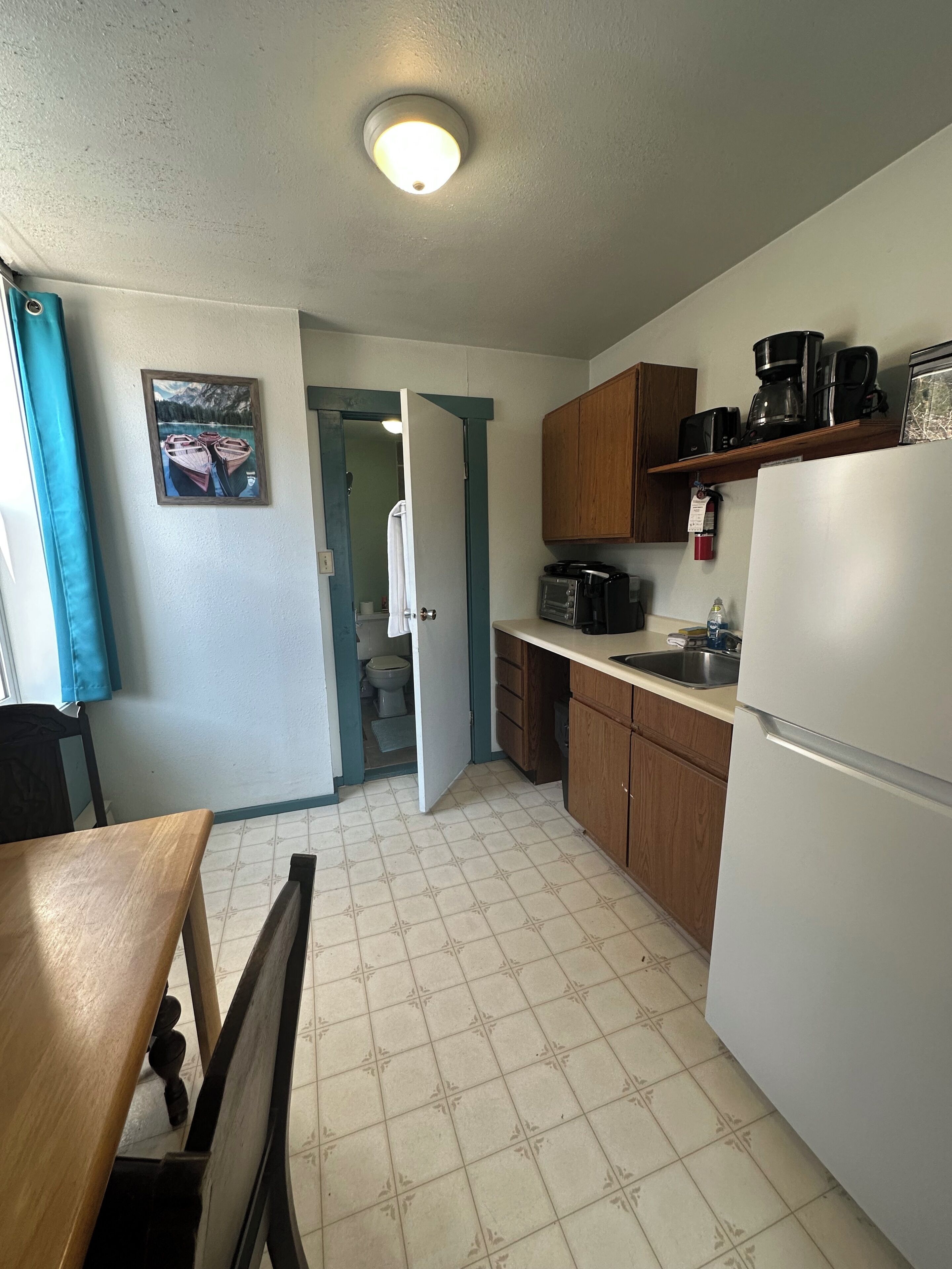 A kitchen with a refrigerator, microwave, and coffee maker. There is a door leading to a bathroom.