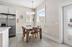 A modern kitchen with a stainless steel refrigerator, wooden dining table, and chairs. There are two windows providing natural light and a view of the outside. - Great Location | Shared Heated Pool | Pet-Friendly (Port Aransas)