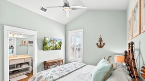 Ein Schlafzimmer mit Bett, Schreibtisch, Stuhl, Deckenventilator, Fenster mit Rollläden und einem an der Wand befestigten Fernseher.