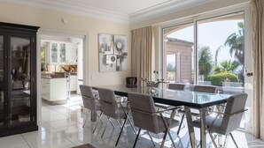 A modern dining area with a glass table and grey chairs, a kitchen in the background, and a view of palm trees through large windows. - Charming 6-bedroom villa (Mouans-Sartoux)