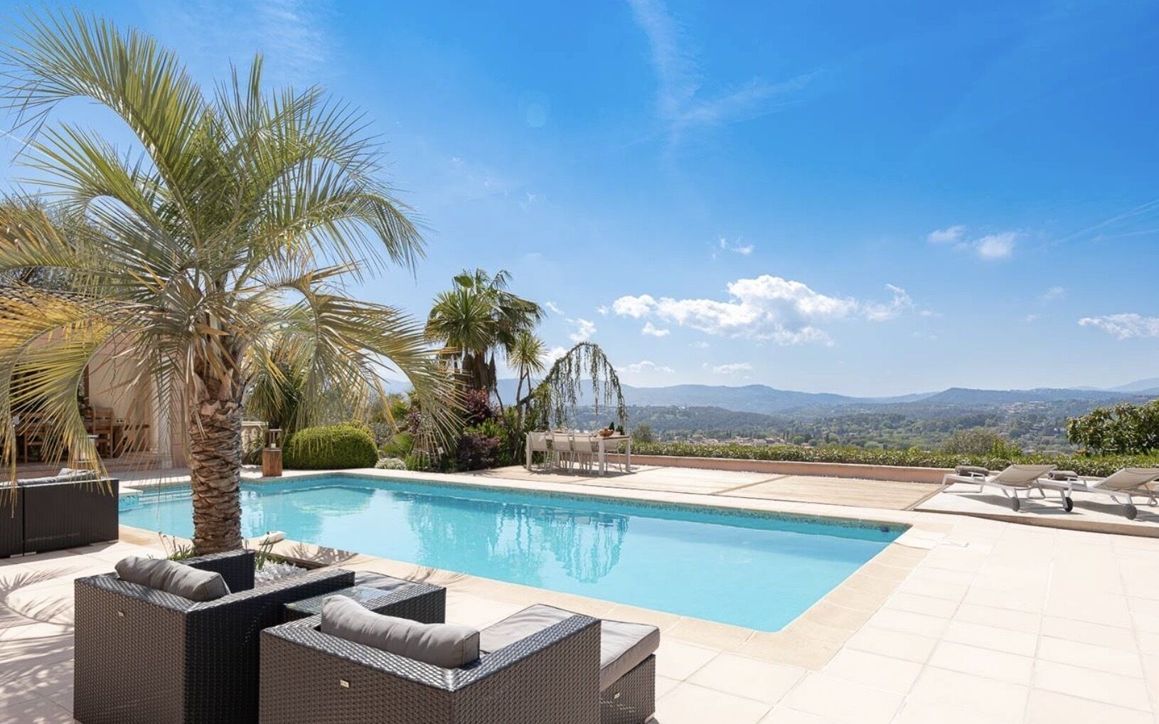 A swimming pool with lounge chairs and a pergola, surrounded by palm trees and a view of distant mountains.