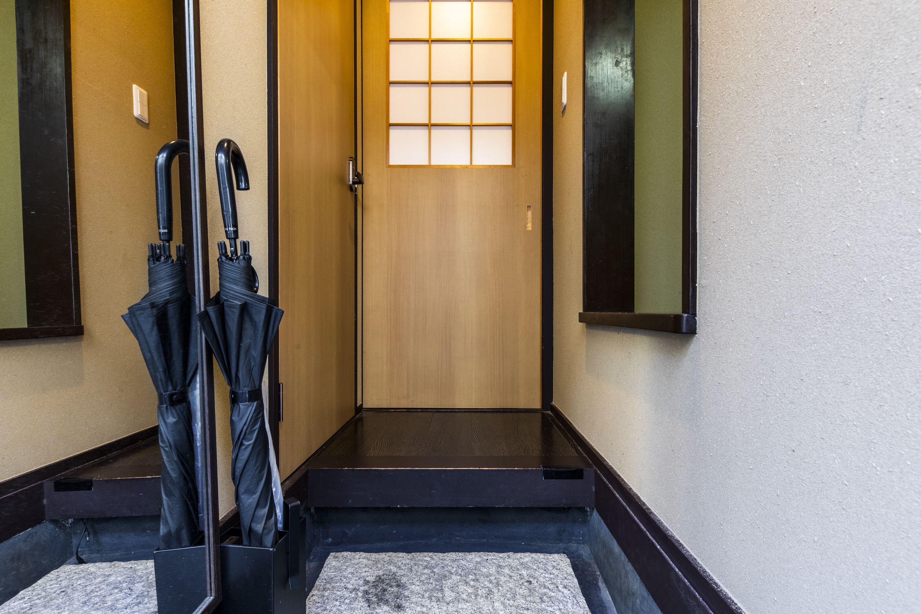 A wooden door with a glass panel, a black metal bench, and two umbrellas leaning against a wall.