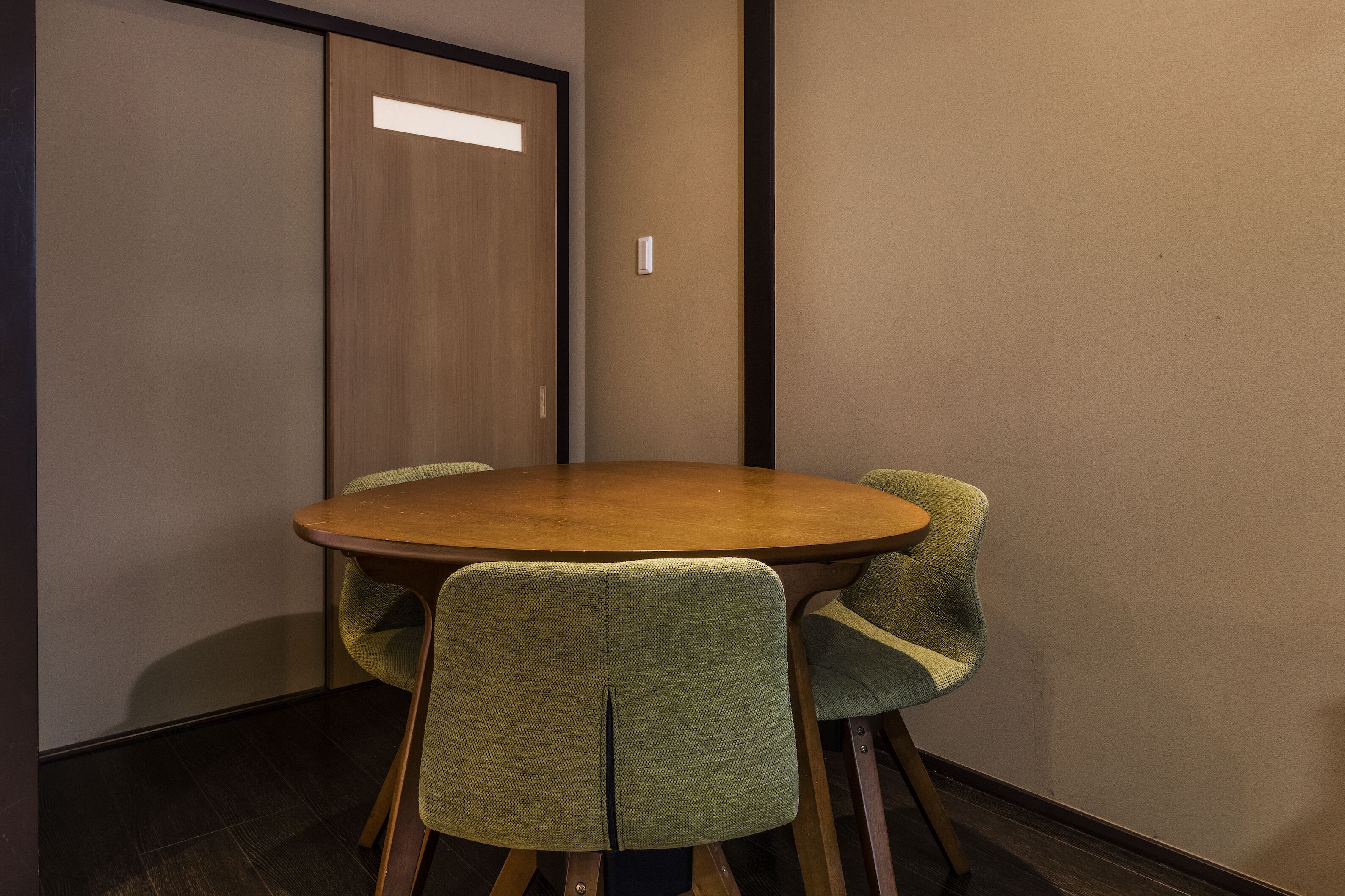 A conference room with a round table and four chairs, a wooden door, and a light switch on the wall.