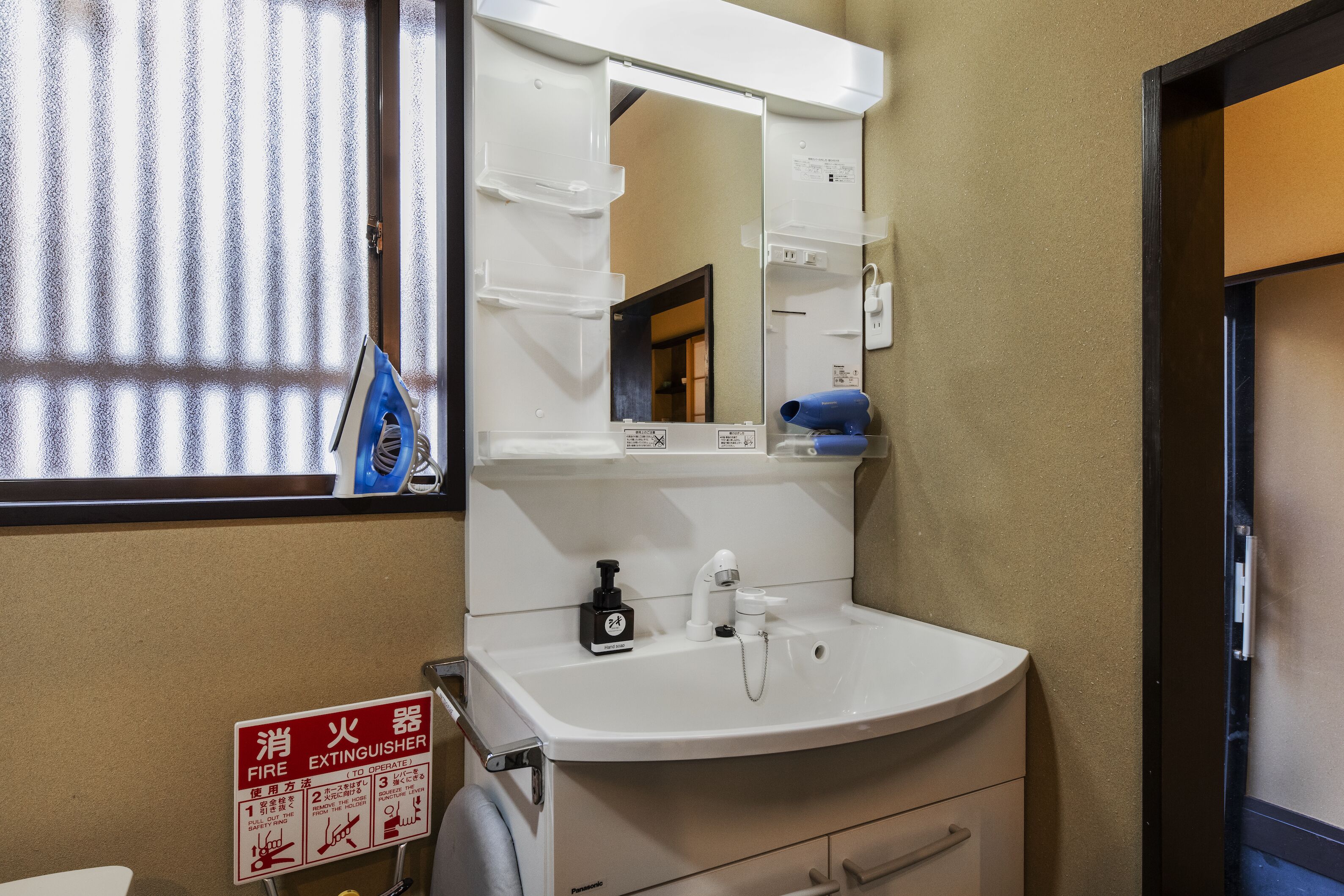 Un baño con lavamanos blanco, un espejo, el cartel de extintor y una ventana con persianas.