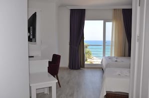 A hotel room with a bed, a desk, a chair, and a TV. There is a balcony with curtains and a view of the sea. - Tepe Beach Hotel (Erdemli)