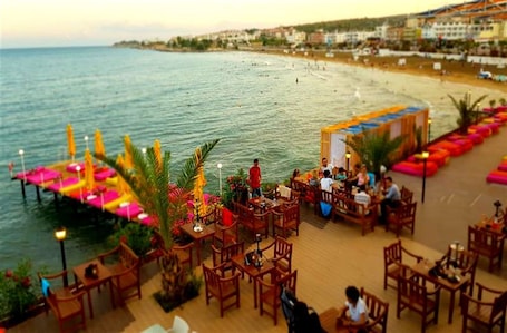 A seaside restaurant with outdoor seating overlooking the ocean, featuring wooden tables and chairs, and a view of the beach and city in the background.