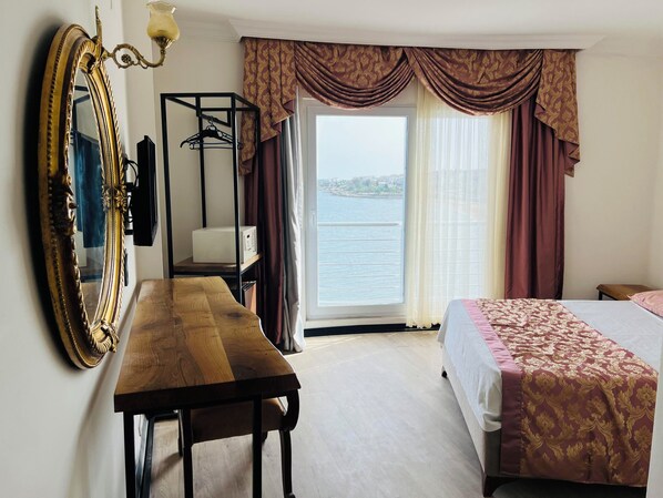 A hotel room with a wooden desk, a bed with a patterned bedspread, a large mirror, and a view of a body of water through a window with curtains. - Tepe Beach Hotel (Erdemli)