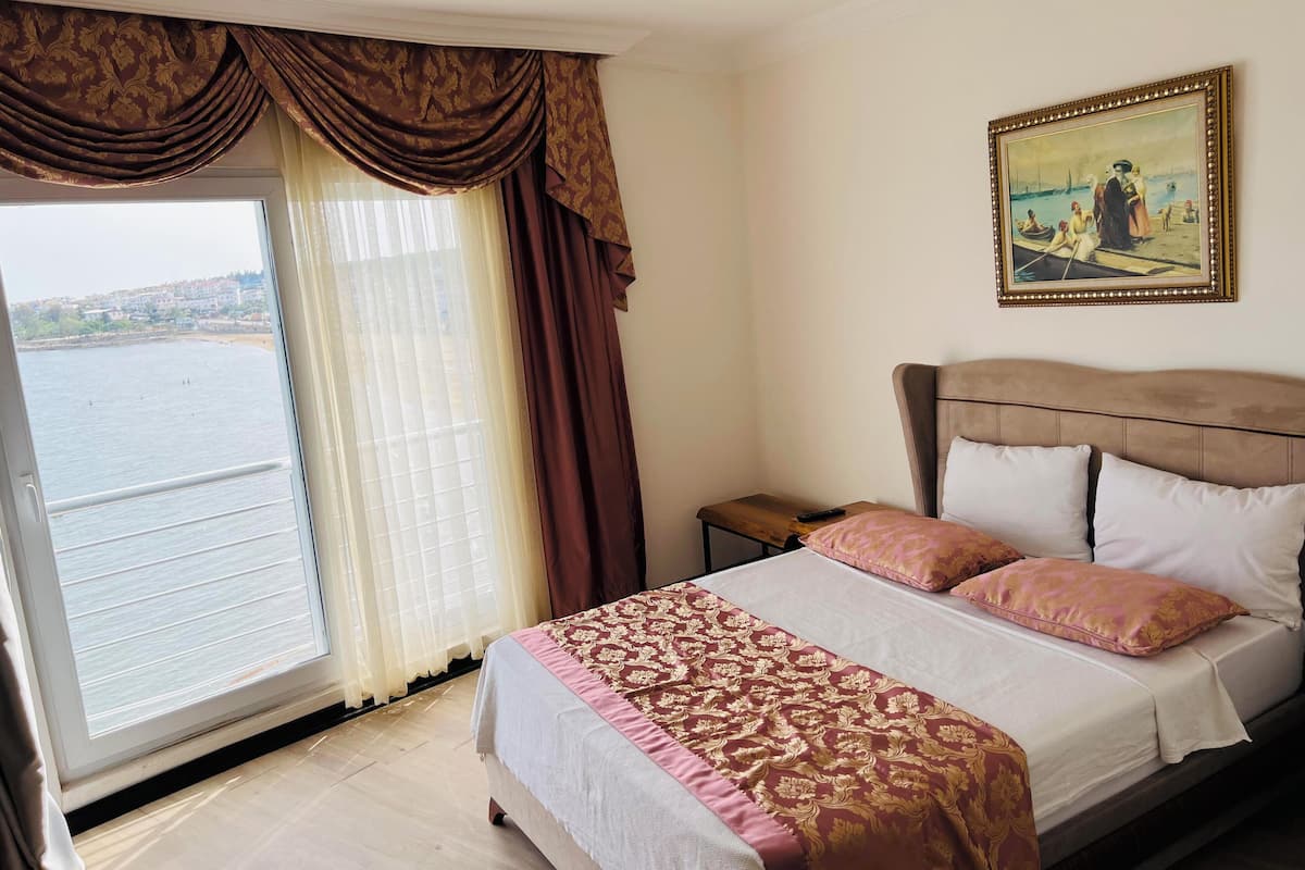 A hotel room with a bed, a nightstand, a painting, and a view of the sea through a window.