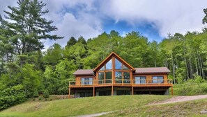 A two-story wooden cabin with large windows and a balcony, surrounded by dense forest. - Hunter Mountain Luxury Cabin | Hot Tub | Fire Pit (Hunter)