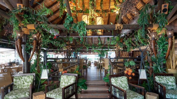 Een restaurant met een houten trap, groene bekleden stoelen en hangende planten.