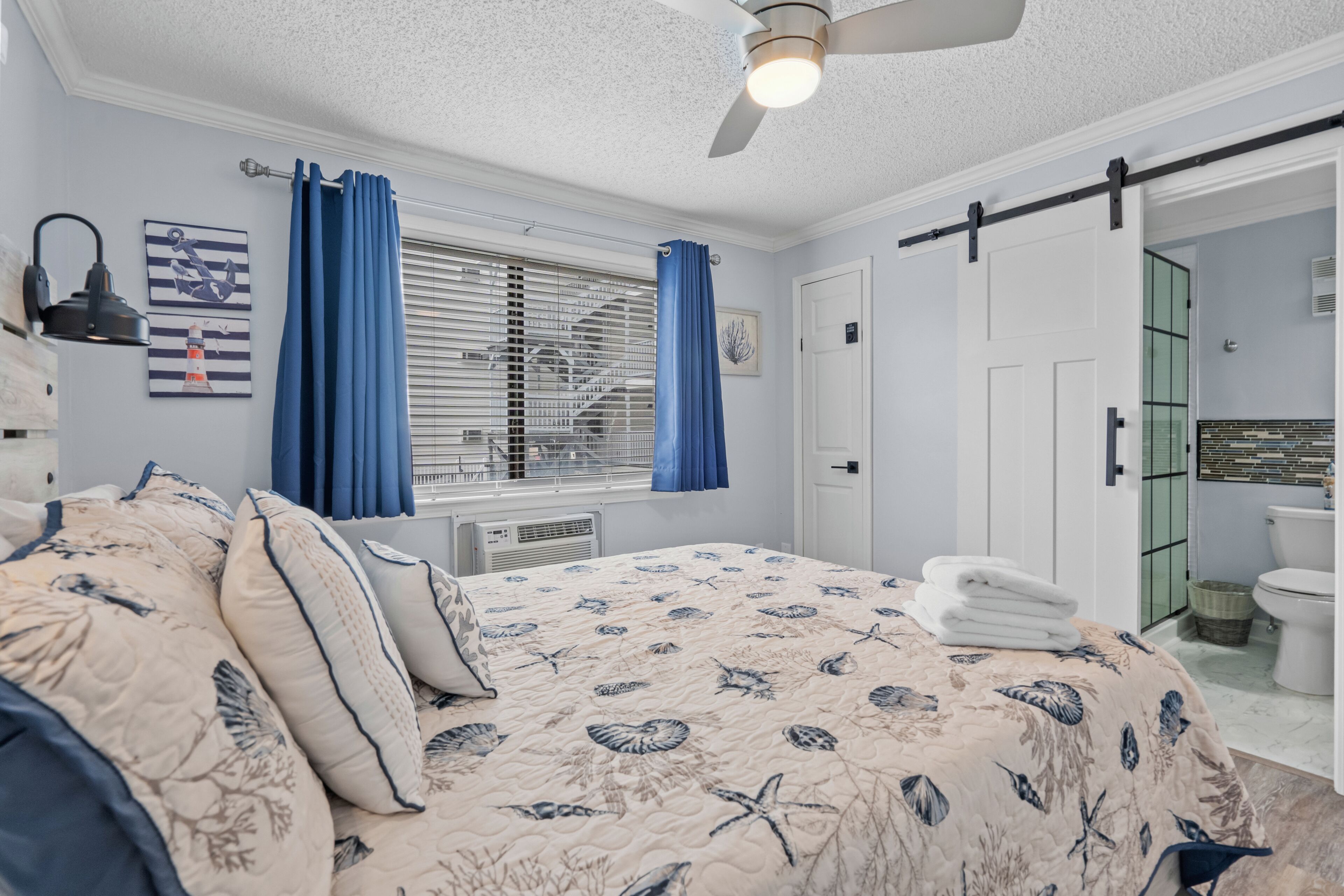 Un dormitorio con cama, ventilador de techo, ventana con persianas, baño con inodoro y puerta que da a otra habitación.