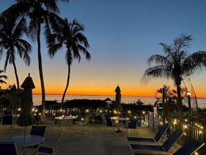 Um terraço à beira-mar com espreguiçadeiras e guarda-sóis, com vista para o oceano ao pôr do sol.