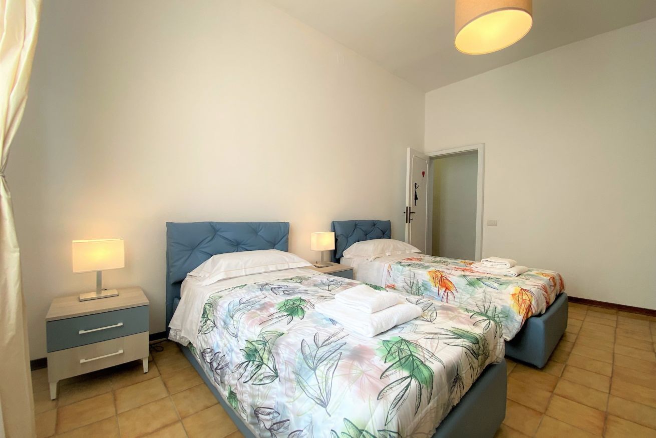 Quarto com duas camas, cada uma com cabeceira azul, mesa de cabeceira com abajur, e uma porta ao fundo.