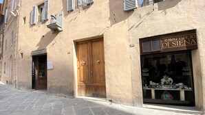A street with a building featuring a wooden door and a storefront with a sign reading "CRAVATTIFICIO DI SIENA". - PALAZZETTO SAN GIOVANNI-in front of the Baptistry (Siena)