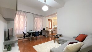 A modern living room with a sofa, a dining table, and chairs. There is a TV on a stand, a small table with a lamp, and a kitchen area in the background. - PALAZZETTO SAN GIOVANNI-in front of the Baptistry (Siena)