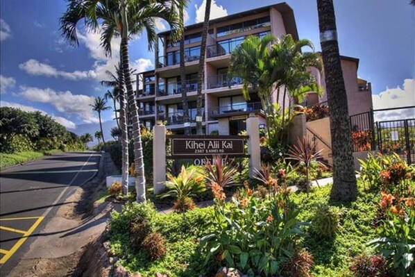 A multi-story hotel with a sign reading "Kihei Alii Kai" and a landscaped garden with tropical plants.