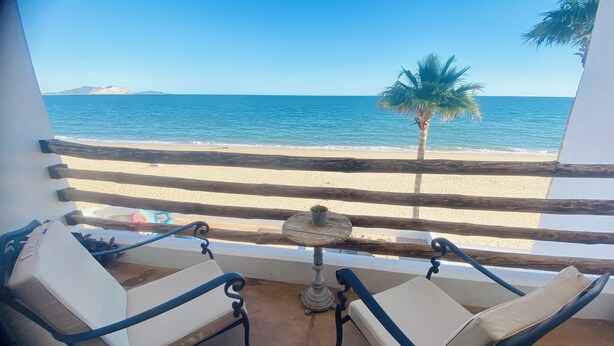 Un balcón con dos sillas acolchadas y una mesita, con vista a una playa con una palmera y el mar.