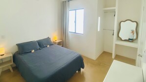 Un dormitorio con cama, lámparas de noche, ventilador de techo, ventana con cortinas, espejo y armario blanco.