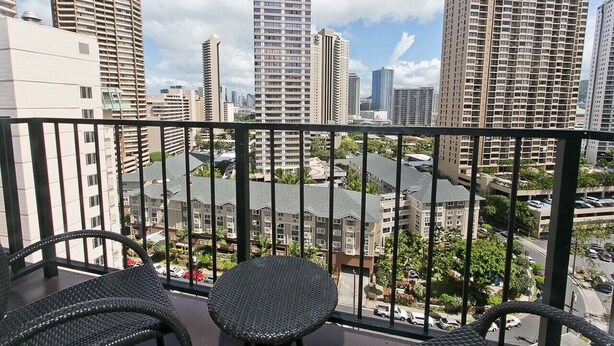 Un balcone con sedie in vimini e vista su palazzi alti e un'area parcheggio.