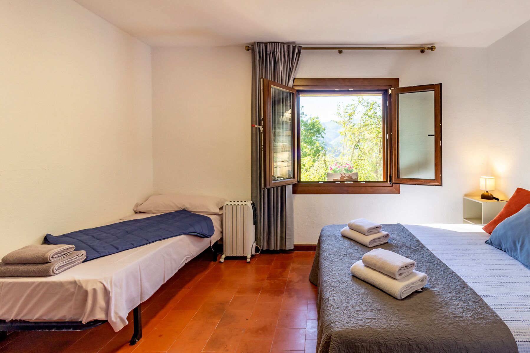 A room with two beds, a window with a view of greenery, a radiator, and a bedside table with a lamp.