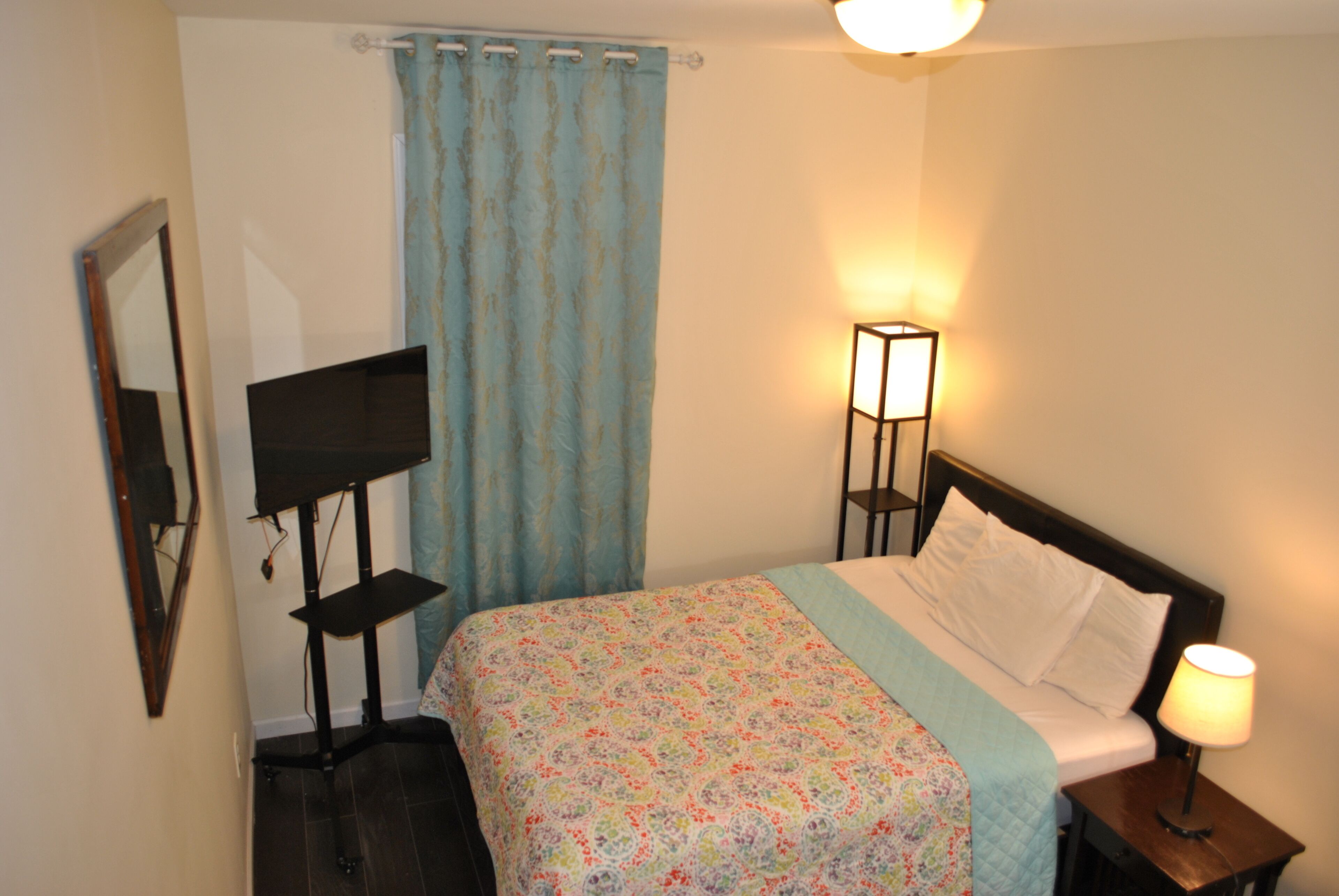 A bedroom with a bed, a nightstand with a lamp, a TV on a stand, and a curtain.