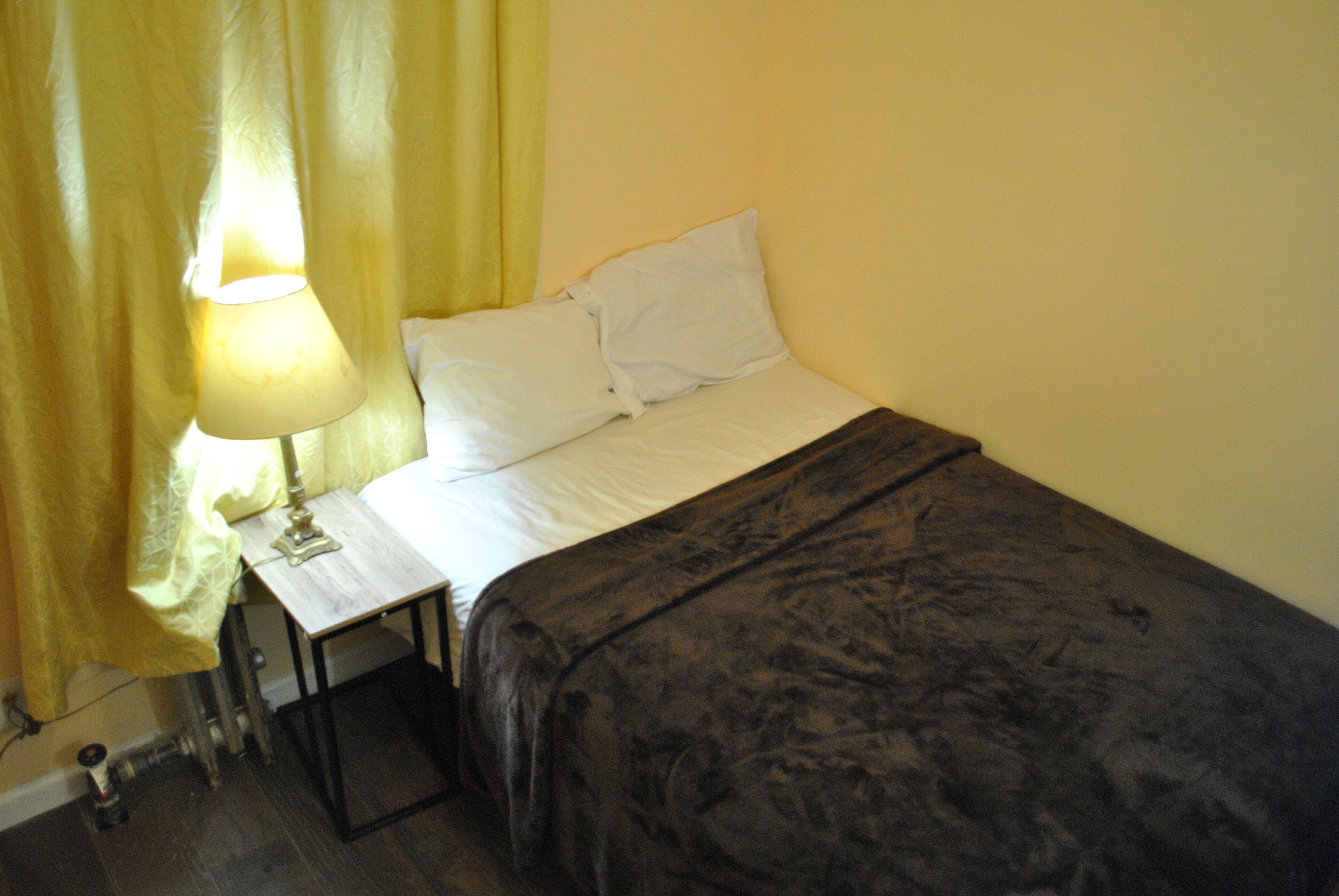 Un lit avec du linge de lit blanc et une couverture sombre, une table de chevet avec une lampe, et un rideau jaune.