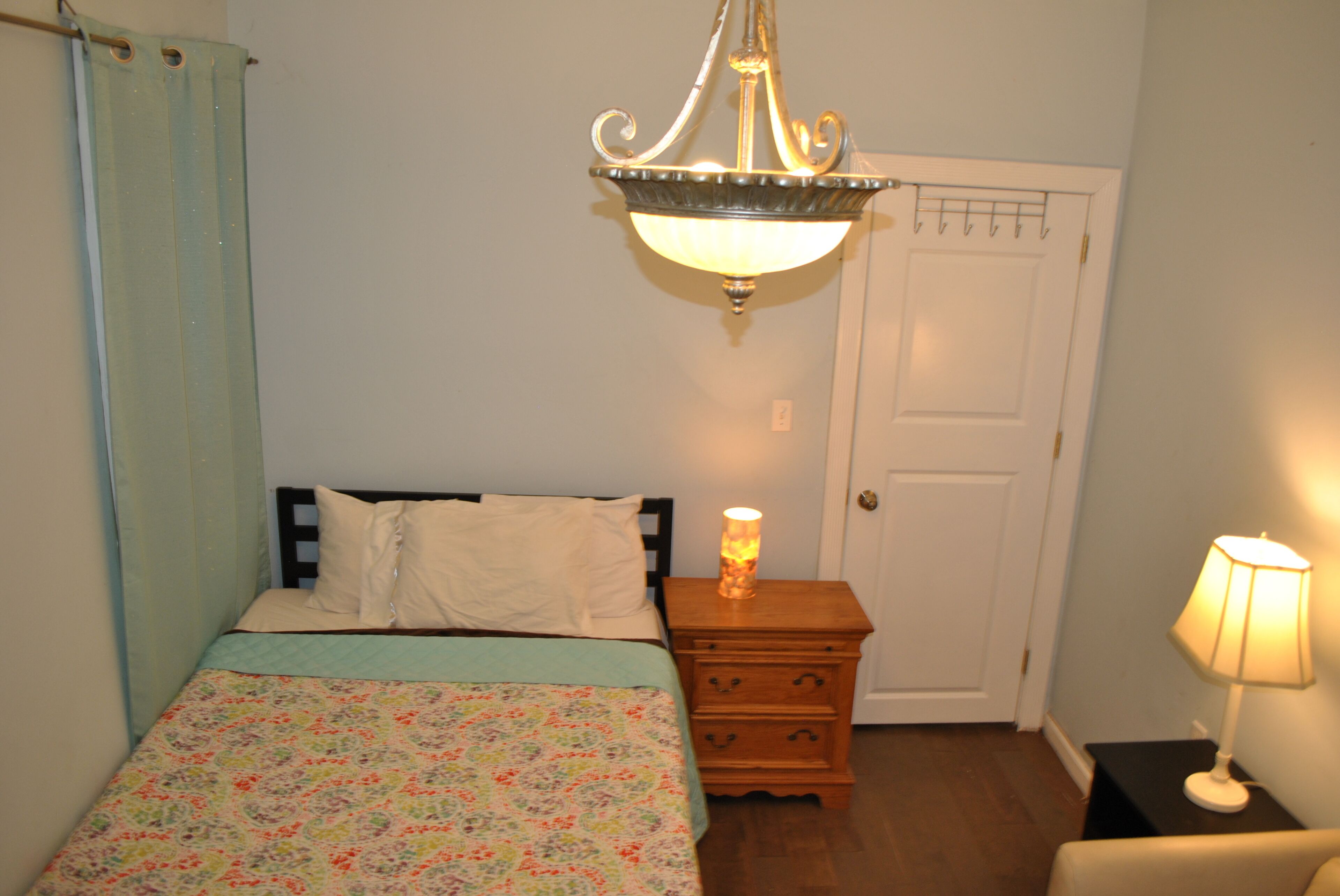 A bedroom with a bed, a nightstand with a lamp, a door, and a hanging light fixture.