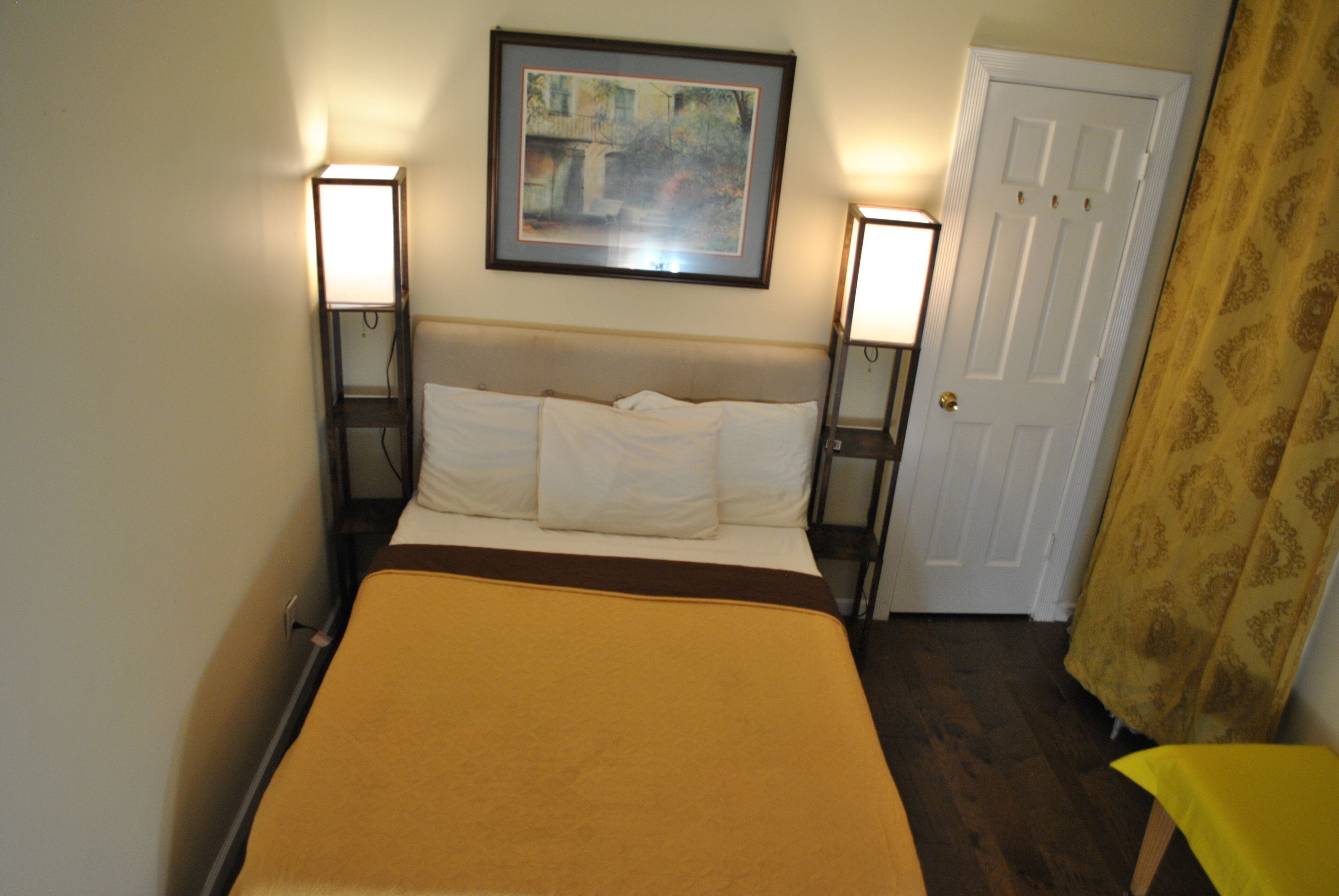 Quarto de hotel com uma cama, duas luminárias de cabeceira, uma porta e um quadro pendurado na parede.