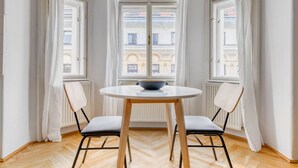 Une pièce d’angle avec une table ronde, deux chaises, un bol posé sur la table et de grandes fenêtres avec des rideaux.