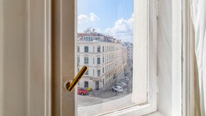 Une vue à travers la fenêtre d’un immeuble à plusieurs étages, donnant sur une rue bordée de voitures garées et un ciel dégagé.