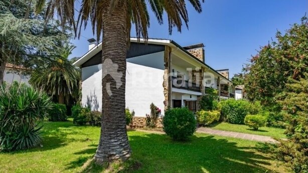 Una casa moderna de dos pisos con un balcón, rodeada por un jardín bien cuidado con diversas plantas y una palmera.