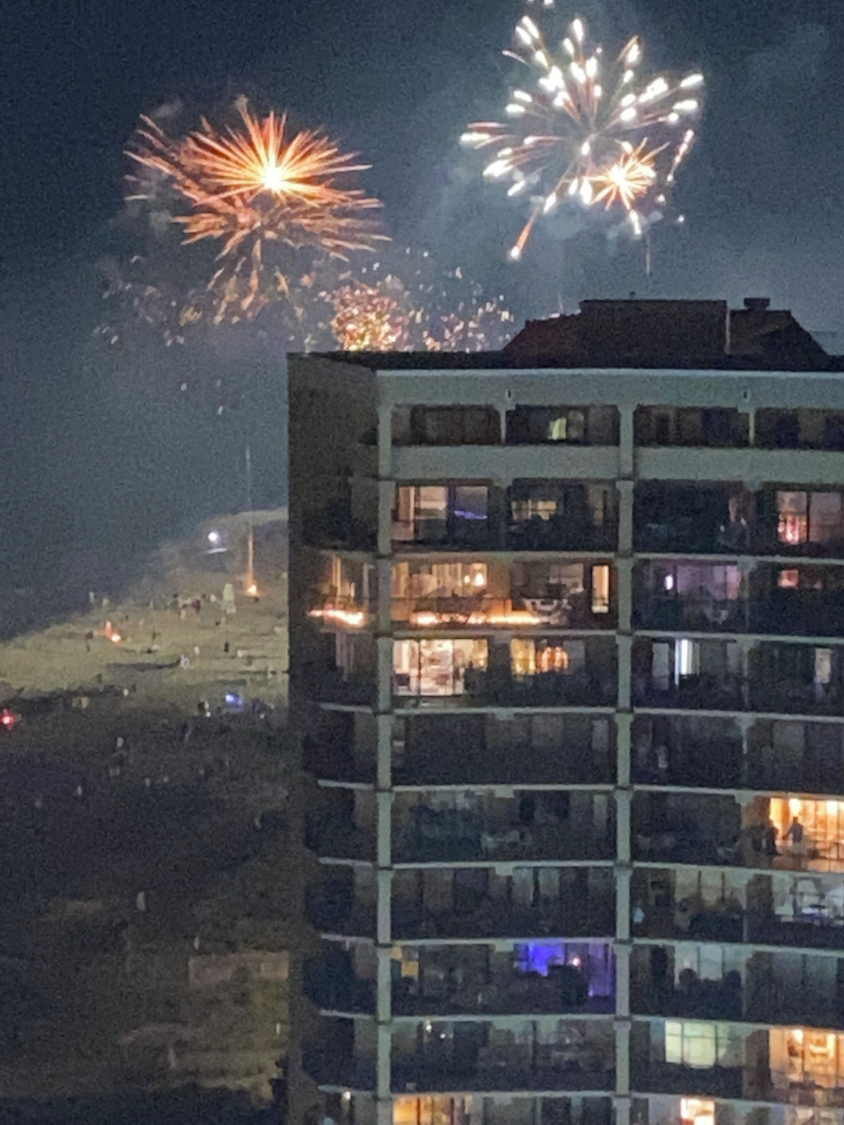 A multi-story building with balconies, a flat roof, and a view of fireworks in the background.