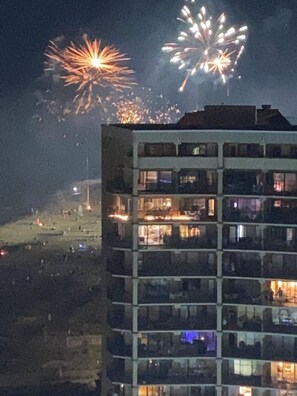 A multi-story building with balconies, a flat roof, and a view of fireworks in the background.