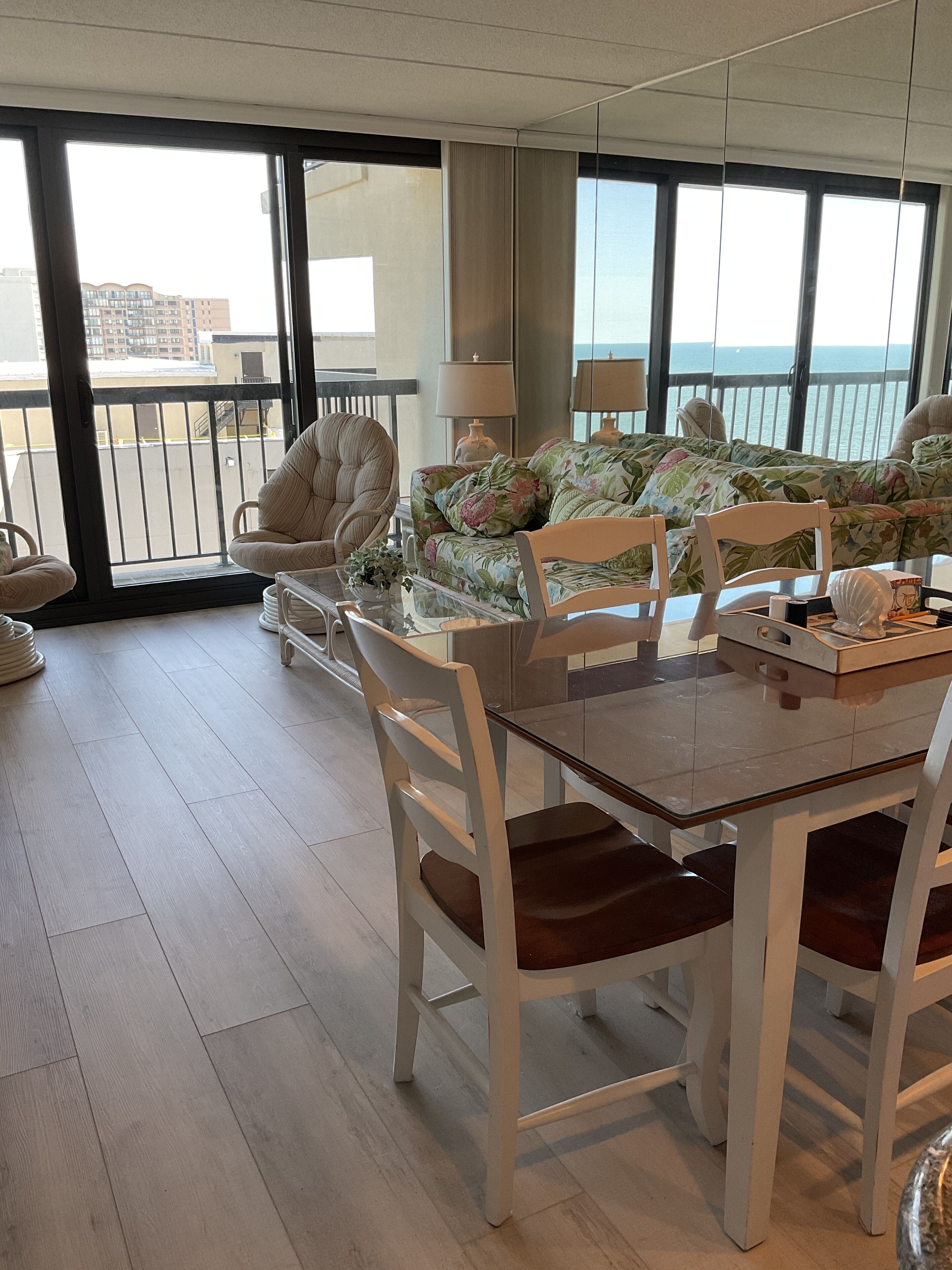 A spacious room with a large window offering a view of the sea. There is a dining table with chairs, a sofa with floral upholstery, and a chair with a cushioned seat.