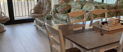 A spacious room with a large window offering a view of the sea. There is a dining table with chairs, a sofa with floral upholstery, and a chair with a cushioned seat.