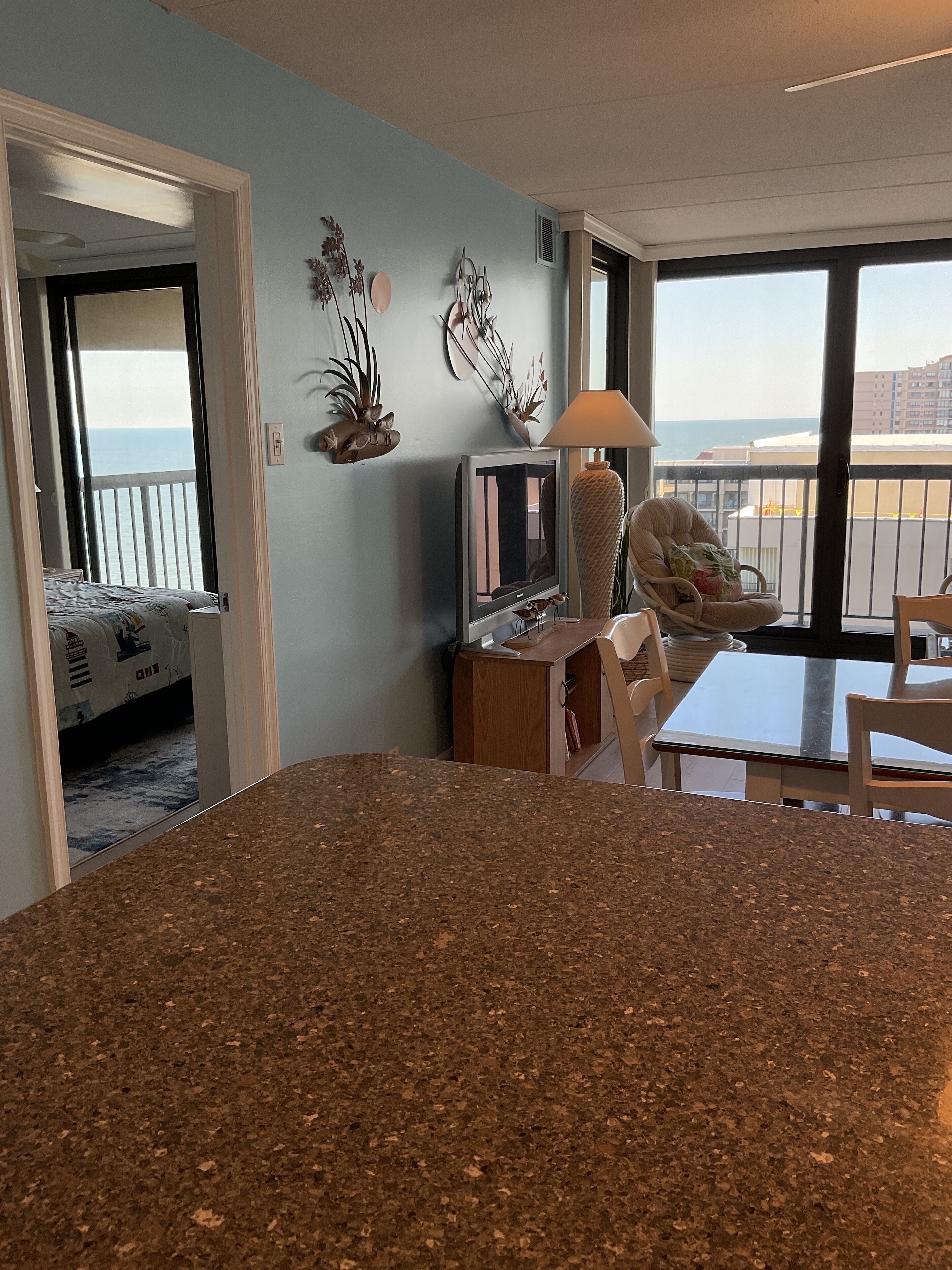 A modern kitchen with granite countertops, a dining area with a table and chairs, a television, and decorative wall art. A balcony with a view of the sea is visible through the window.