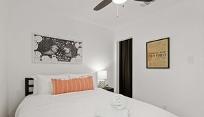 Een netjes opgemaakt bed met witte en gestreepte kussens, een plafondventilator, een ingelijste poster aan de muur en een klein nachtkastje met een lamp.