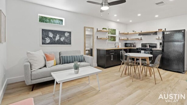 Un salon moderne avec un canapé gris, une table basse blanche et un coin repas comprenant une table ronde et des chaises. On y trouve également une cuisine avec des meubles noirs et un réfrigérateur noir.