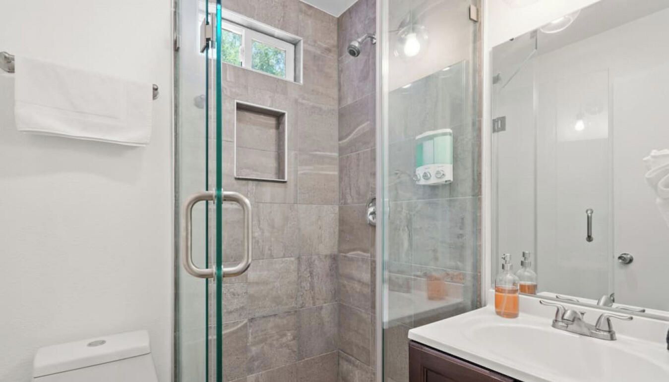 A modern bathroom with a glass-enclosed shower, a white sink with a mirror, and a small window above the shower.