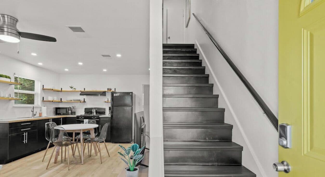 A modern kitchen with a dining area, black staircase, and a bright yellow door.
